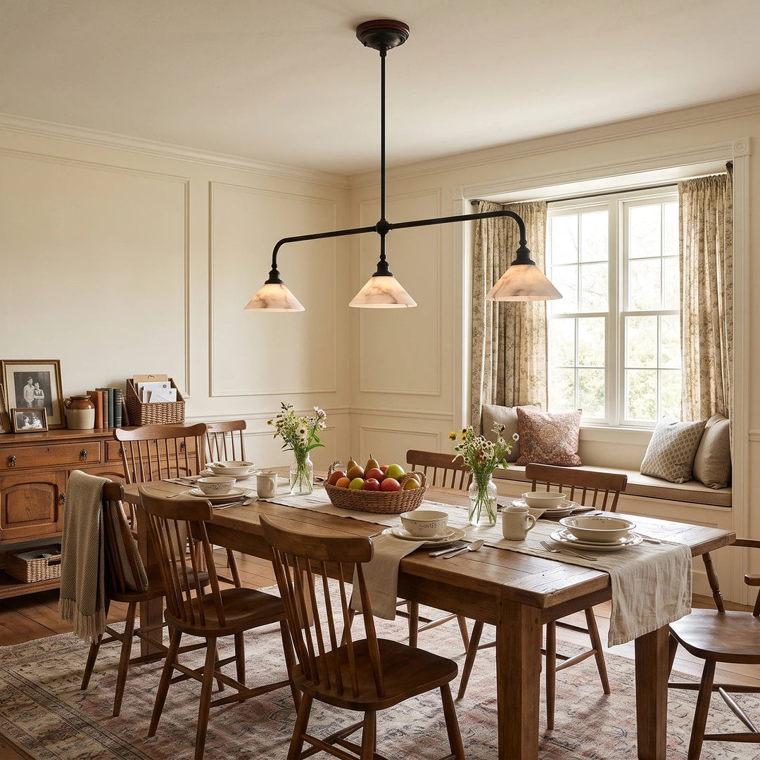 A three-light black metal bar pendant chandelier with conical shades hangs over a wooden dining table set for a meal in a warm, classic dining room with wood chairs, a window with patterned curtains, and a built-in window seat.
