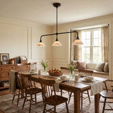 A three-light black metal bar pendant chandelier with conical shades hangs over a wooden dining table set for a meal in a warm, classic dining room with wood chairs, a window with patterned curtains, and a built-in window seat.