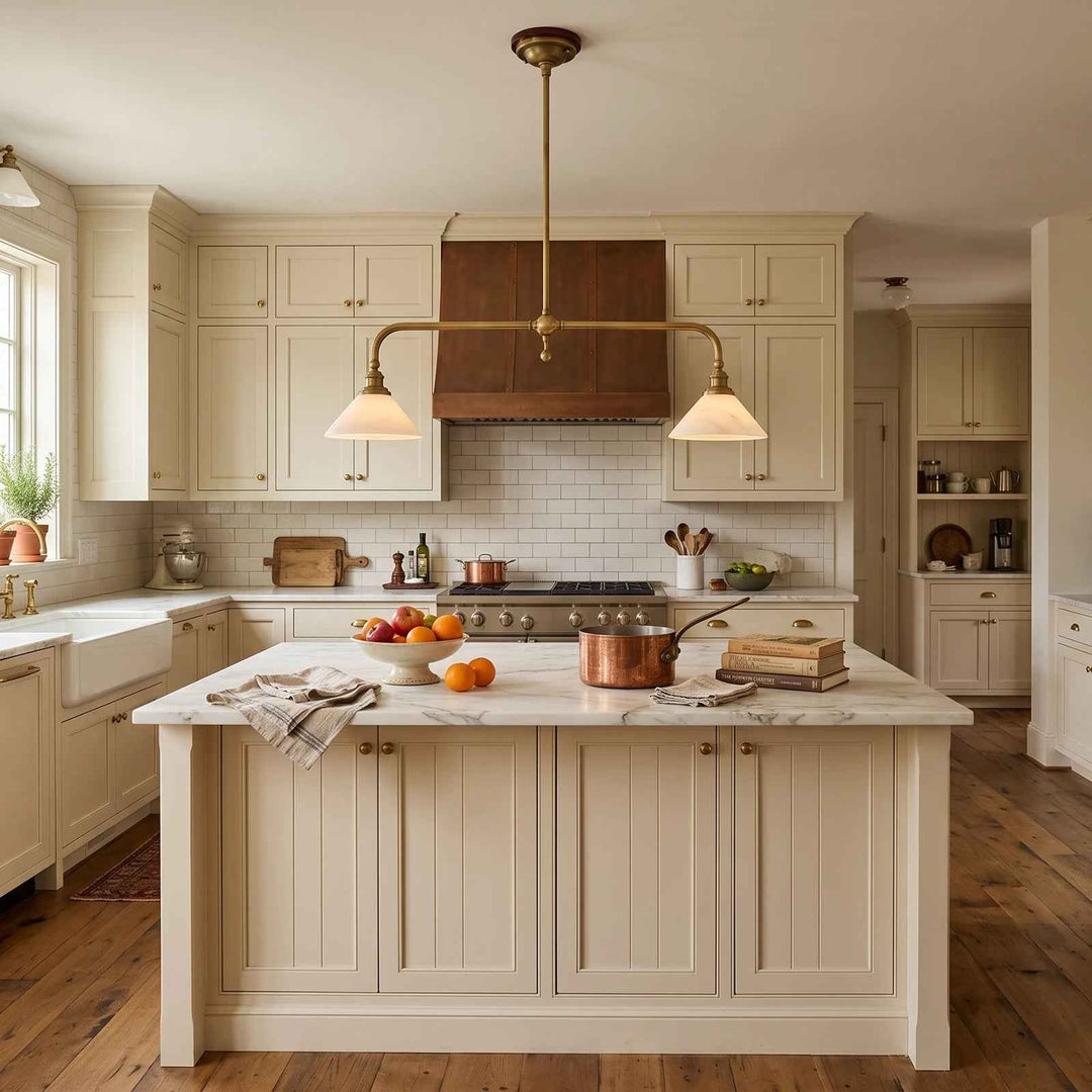 A bright, traditional kitchen interior with cream cabinetry, a white subway tile backsplash, a dark wood range hood, and a central island with a marble countertop. A brass double-arm pendant light hangs above the island, and copper cookware sits on the counter.