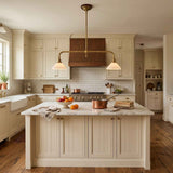 A bright, traditional kitchen interior with cream cabinetry, a white subway tile backsplash, a dark wood range hood, and a central island with a marble countertop. A brass double-arm pendant light hangs above the island, and copper cookware sits on the counter.