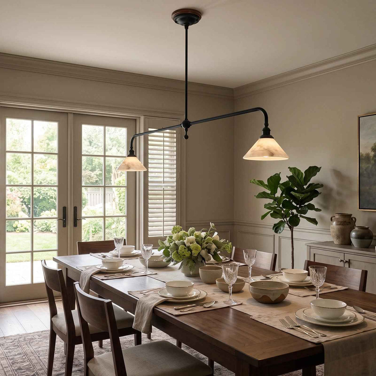 A modern black metal multi-arm pendant light with two warm white conical shades hangs over a wooden dining table set for a meal in a tastefully decorated dining room.