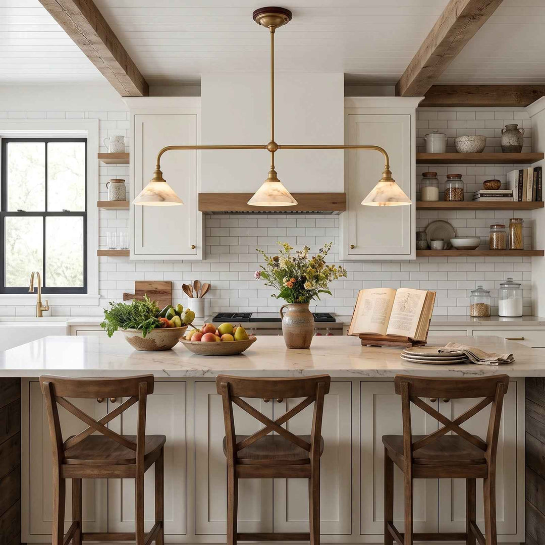 A modern kitchen interior with white cabinetry, a marble island, three wooden bar stools, a brass three-light pendant chandelier, exposed wood ceiling beams, and a white subway tile backsplash.