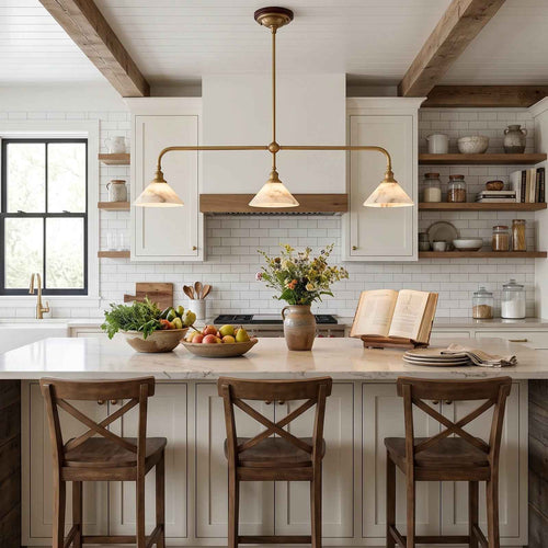 A modern kitchen interior with white cabinetry, a marble island, three wooden bar stools, a brass three-light pendant chandelier, exposed wood ceiling beams, and a white subway tile backsplash.