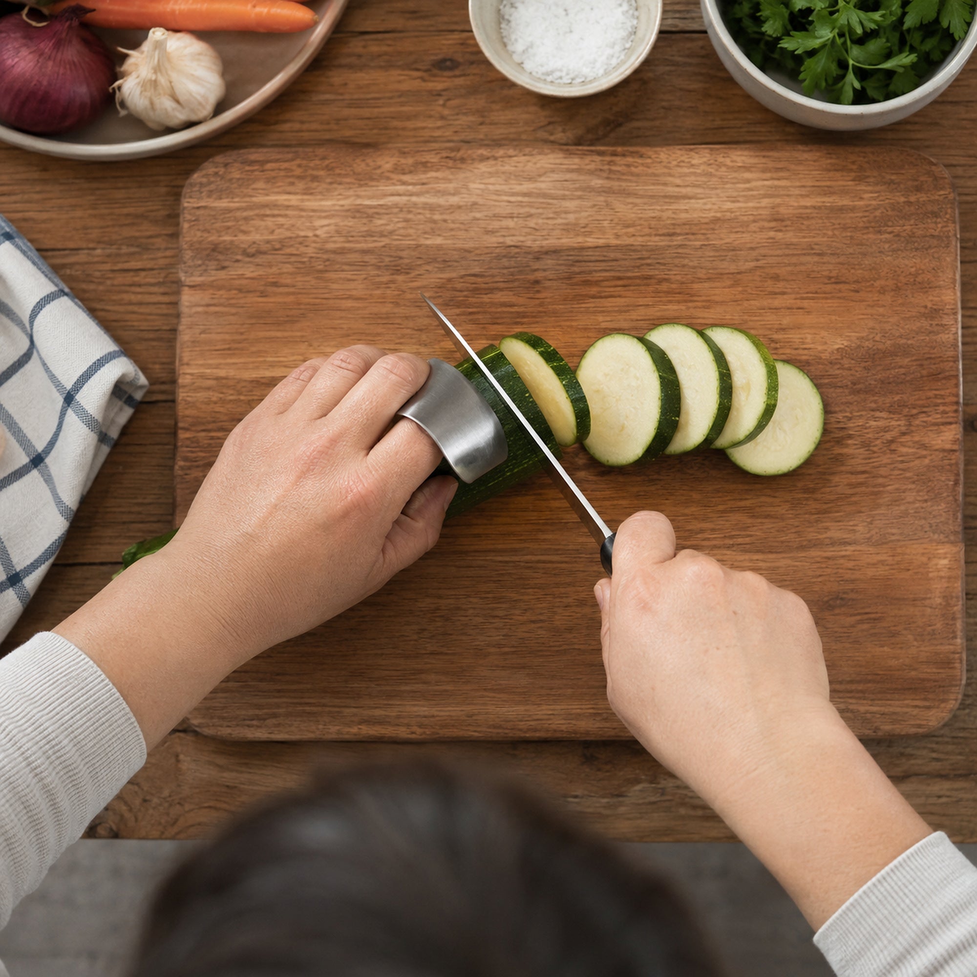 Top-down view of a cook using the stainless steel finger guard to slice zucchini on a wooden board