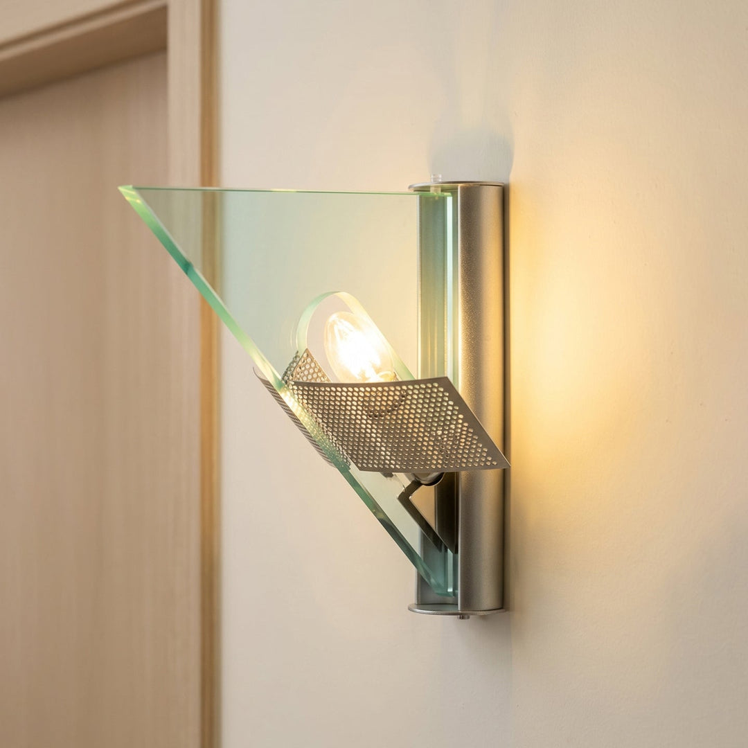 Illuminated silver wall sconce with triangular clear glass and perforated metal accents on a beige wall beside decorative trim.