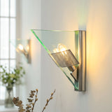 Silver wall sconce with triangular glass casts warm light on a white wall, with another sconce and window behind.