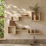 Light natural wood floating shelves mounted on a textured beige wall in a kitchen setting, styled with a ceramic vase, greenery, a wooden cutting board, plates, and mugs. A window on the left casts dappled sunlight across the scene.