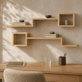 Light natural wood floating wall shelves arranged in an L-shape above a wooden desk, styled with books, small bowls, and a cup. A textured beige chair is partially visible in the foreground against a warm beige wall with soft leaf shadows.
