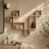 Light natural wood floating wall shelves arranged decoratively on a beige wall above a stone sink, with rolled towels, a small vase, a candle, and an amber bottle displayed in the shelves. A mirror, wall sconce, and plant leaves are visible in a softly lit bathroom setting.