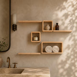 Light natural wood floating wall shelves mounted on a textured beige wall in a bathroom setting, styled with a candle, small vase, bottle, and rolled towels.