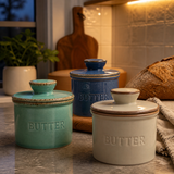 Three ceramic butter canisters with lids in teal, blue, and cream are arranged on a kitchen counter with a loaf of bread, cutting board, and tiled backsplash in the background.