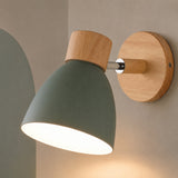 A modern wall-mounted light fixture with a matte sage green cone-shaped shade, natural wood accents, and a round wooden backplate, shown lit against a warm neutral wall.
