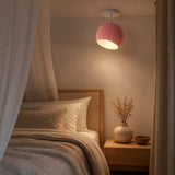 A modern ceiling light with a pink spherical shade and white canopy, glowing warmly in a bedroom setting next to a bed with a wooden headboard and neutral bedding.
