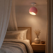 A modern ceiling light with a pink spherical shade and white canopy, glowing warmly in a bedroom setting next to a bed with a wooden headboard and neutral bedding.