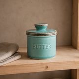 A turquoise ceramic butter jar with a matching lid and the word 'BUTTER' embossed on the front, displayed on a wooden shelf next to a folded beige cloth.