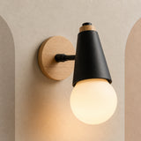 A modern wall-mounted light fixture with a black cone-shaped shade, a warm white globe bulb, a light wood circular backplate, and a small wood accent ring near the top, shown lit against a beige textured wall.