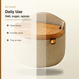 A ceramic salt cellar with a speckled beige body and a light brown wooden lid, shown on a clean off-white background with text listing features like 'Open scoop', 'Counter ready', and 'Tray on sets'.