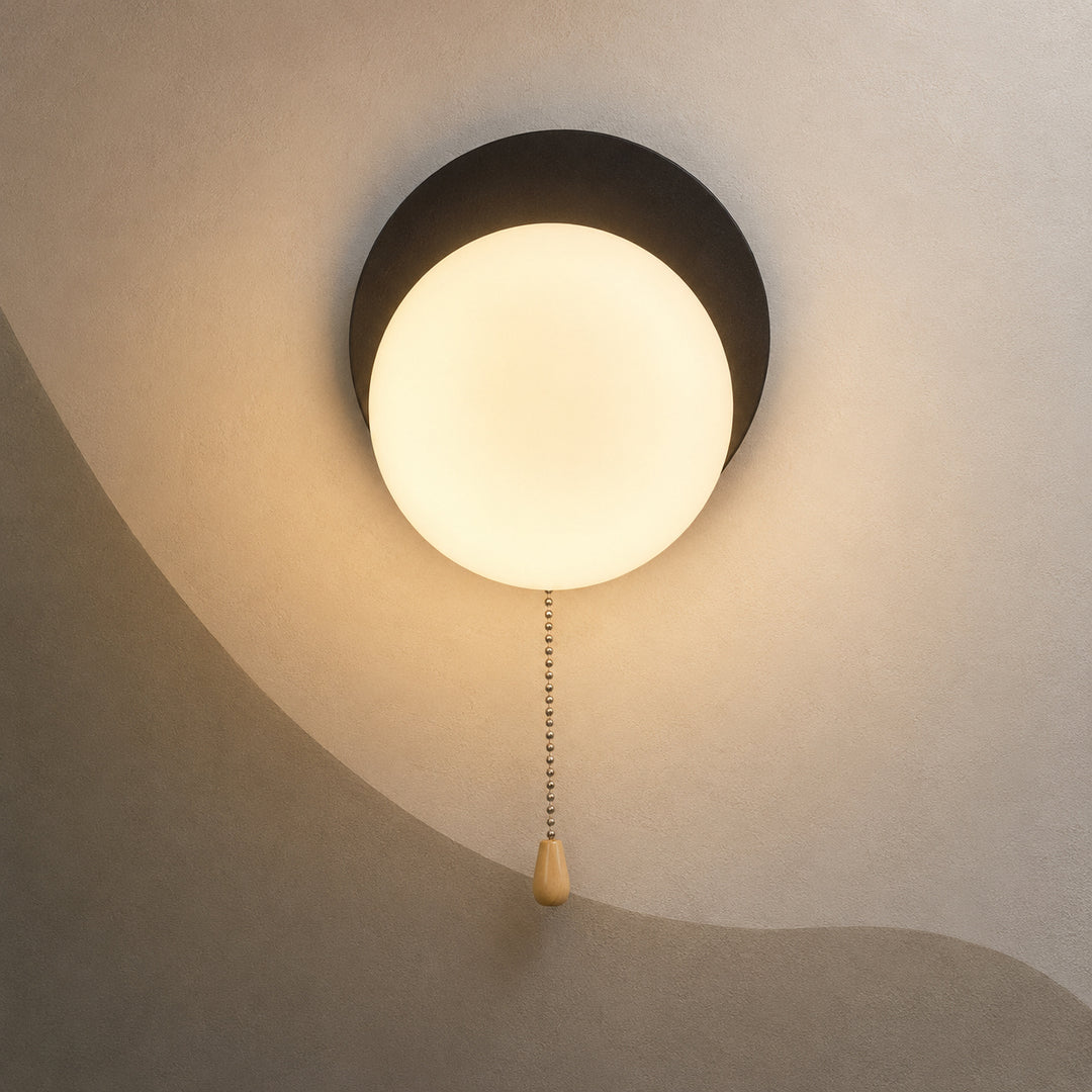 A modern ceiling light fixture with a glowing frosted white globe and a matte black circular mount, shown installed on a textured beige ceiling with a pull chain.