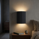 A modern black wall sconce with vertical fluted detailing, mounted on a textured beige wall above a wooden nightstand. The warm light from the lamp casts a glow onto the wall and illuminates a book and a ceramic cup on the bedside table, with a bed visible on the right.