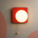 A modern square wall sconce with a vibrant red frame and a glowing white circular light, hanging from a pull chain with a wooden tassel against a textured gray wall with abstract pink shapes.