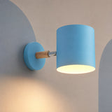 A modern wall-mounted light fixture with a cylindrical blue shade, a round blue wall plate, a natural wood accent, and a silver metal arm, shown lit against a neutral beige wall.