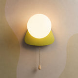 A modern wall sconce with a glowing white spherical bulb and a bright yellow-green conical shade, mounted on a beige wall with abstract wavy patterns. A pull chain with a small wooden knob hangs below.