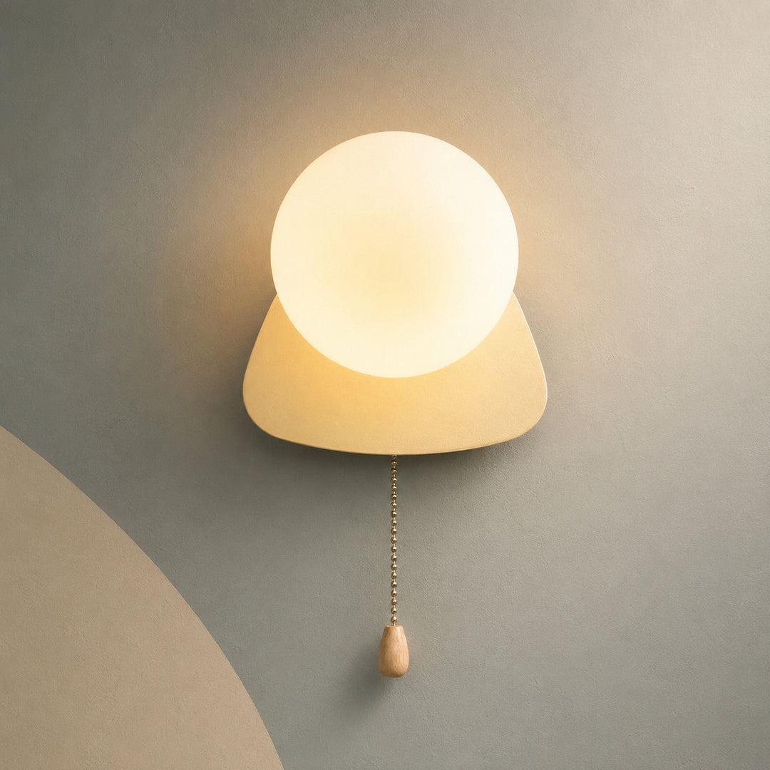 A modern wall-mounted light fixture with a glowing frosted oval globe and a triangular shade, shown against a gray wall with a pull chain and wooden switch below.
