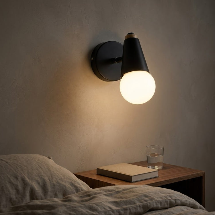 Black globe wall sconce lit at night above a walnut nightstand beside a bed