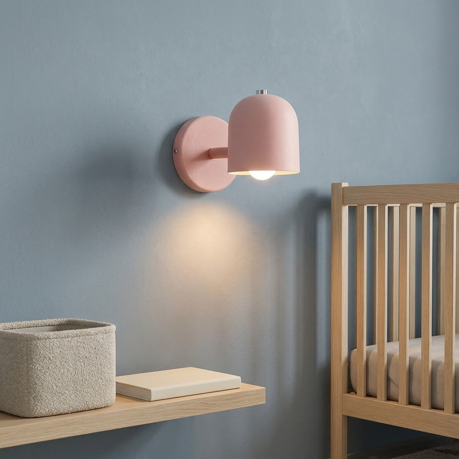 Pink dome wall sconce lit beside a pale oak crib in a nursery corner