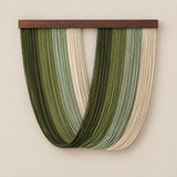 A U-shaped wall hanging made of many vertical strands in shades of olive green, sage, and cream, suspended from a dark wood bar against a warm beige wall.