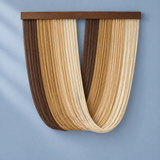 A modern wall-mounted decorative sculpture made of many narrow vertical wood strips in dark brown, tan, and light beige, arranged in a U-shape with a dark wood top bar, shown against a soft blue wall.