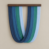 A U-shaped wall hanging made of many vertical strands in shades of blue, teal, and pale blue, suspended from a dark wood bar against a warm beige wall.