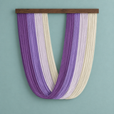 A wall-mounted decorative hanging made of many narrow vertical strands in alternating purple and cream tones, suspended from a dark wooden bar against a pale blue-green wall.