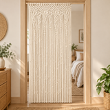 A cream macrame door curtain hanging in a doorway, with a softly lit bedroom visible behind it. The room has light wood trim, a bed with neutral bedding, potted plants, and woven decor.