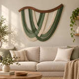 A large woven wall hanging with draped green and cream cords and a curved wooden top, displayed above a light beige sofa in a styled living room with plants and pillows.