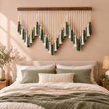A bohemian-style woven wall hanging with green and cream tassels and wooden beads, displayed above a bed with matching green and cream bedding in a warm beige bedroom.