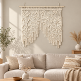 A cream macrame wall hanging with intricate woven patterns and long fringe, suspended from a wooden rod above a light beige sofa in a styled living room.
