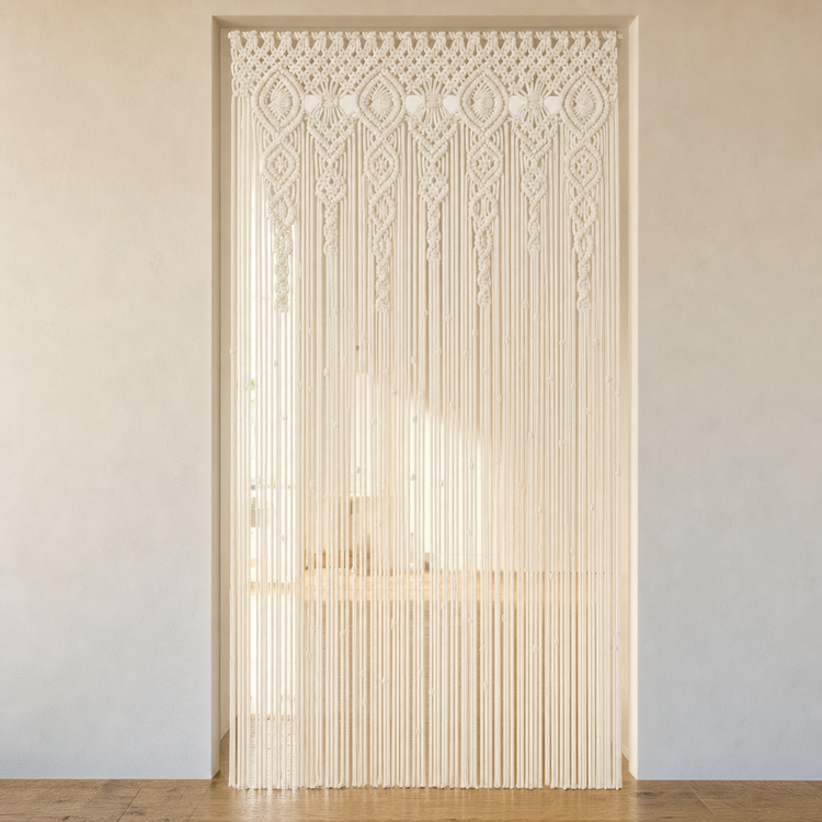 An off-white macrame curtain with intricate woven patterns and long hanging strands, displayed as a door curtain in a minimalist room setting with beige walls and a wooden floor.