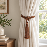 A cream-colored linen curtain panel tied back with a brown braided rope tieback and tassel, hanging next to a window in a styled room.
