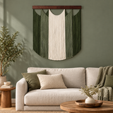 A green and cream macrame wall hanging with fringe details is displayed on a sage green wall above a light beige sofa in a styled living room setting.