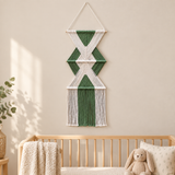A green and white geometric macrame wall hanging displayed above a light wood crib in a softly lit nursery room.