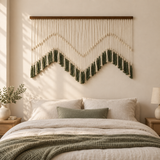 A bohemian-style macrame wall hanging with a mountain-like zigzag pattern in cream and dark green, displayed above a neatly made bed in a softly lit bedroom.