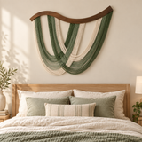 A bohemian-style woven wall hanging with green and cream cords in a harp-like shape, displayed above a wooden bed headboard in a softly lit bedroom.