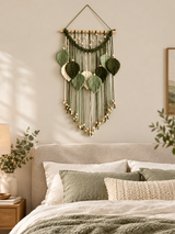 A green leaf macrame wall hanging with wooden beads and a natural wood dowel, displayed on a beige wall above a bed with neutral-toned bedding in a softly lit bedroom.