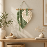 A bohemian-style woven wall hanging made from two leaf-like forms hangs on a wooden dowel on a beige wall above a wooden console table. The left leaf is sage green and the right leaf is cream, with macrame details and hanging beads.