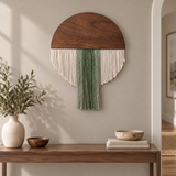 A mushroom-shaped bohemian wall hanging with a dark wood cap, cream fringe, and a green fringe center, displayed on a beige wall above a wooden console table with decorative items.