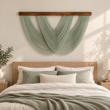 A bohemian-style woven wall hanging with a curved, layered design in sage green, suspended from a dark wood rod above a bed with neutral-toned bedding and pillows.