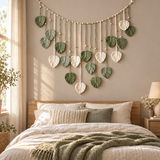 A bohemian-style leaf wall hanging with green and cream leaf-shaped ornaments suspended from a wooden garland, displayed above a bed in a warm, sunlit bedroom.