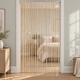 A gold beaded curtain hanging in a doorway, separating a bedroom with white bedding from a sunlit room with light gray walls and a wooden side table.