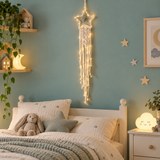 A hanging star-shaped macrame wall decor piece with warm white string lights and tassels, displayed on a blue wall above a child's bed in a styled bedroom.